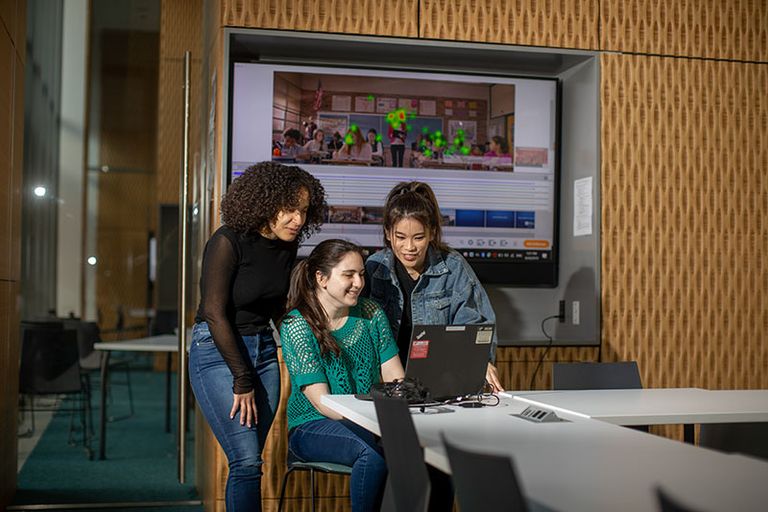 Three women huddle around a laptop.