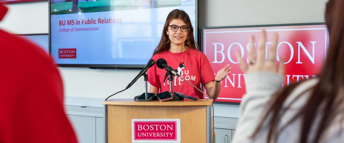 Woman at lectern speaking in front of large tv screen.