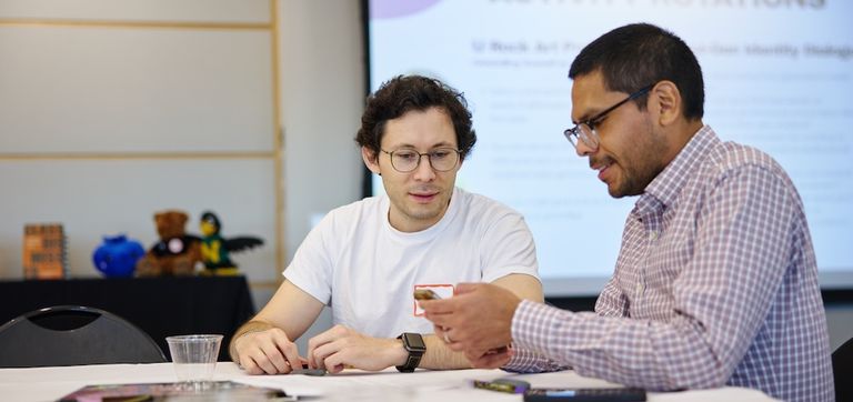 Two men sit at a table looking at a cell phone that one of them is holding. Behind them, a slide from a presentation is projected onto a screen.